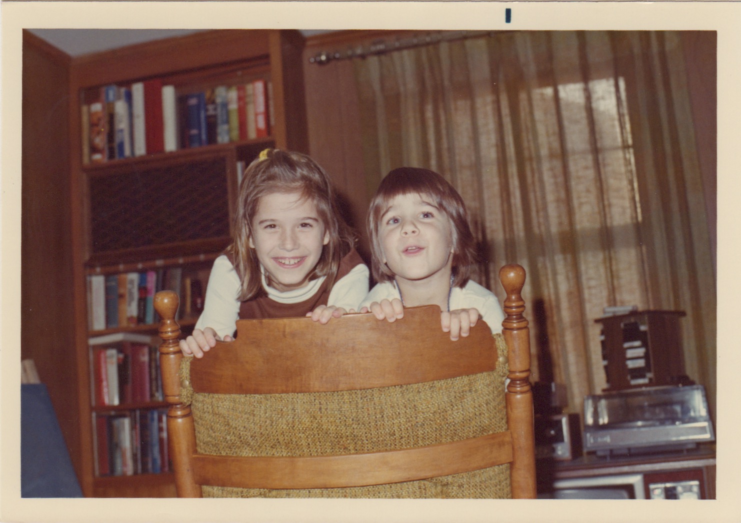 photo of Pam and her sister Bridget standing up looking over the back of a family room chair that has an open wood slat chair back. In the background is a floow-to-ceiling bookcase full of books. Next to is a French door with a curtain for privacy.