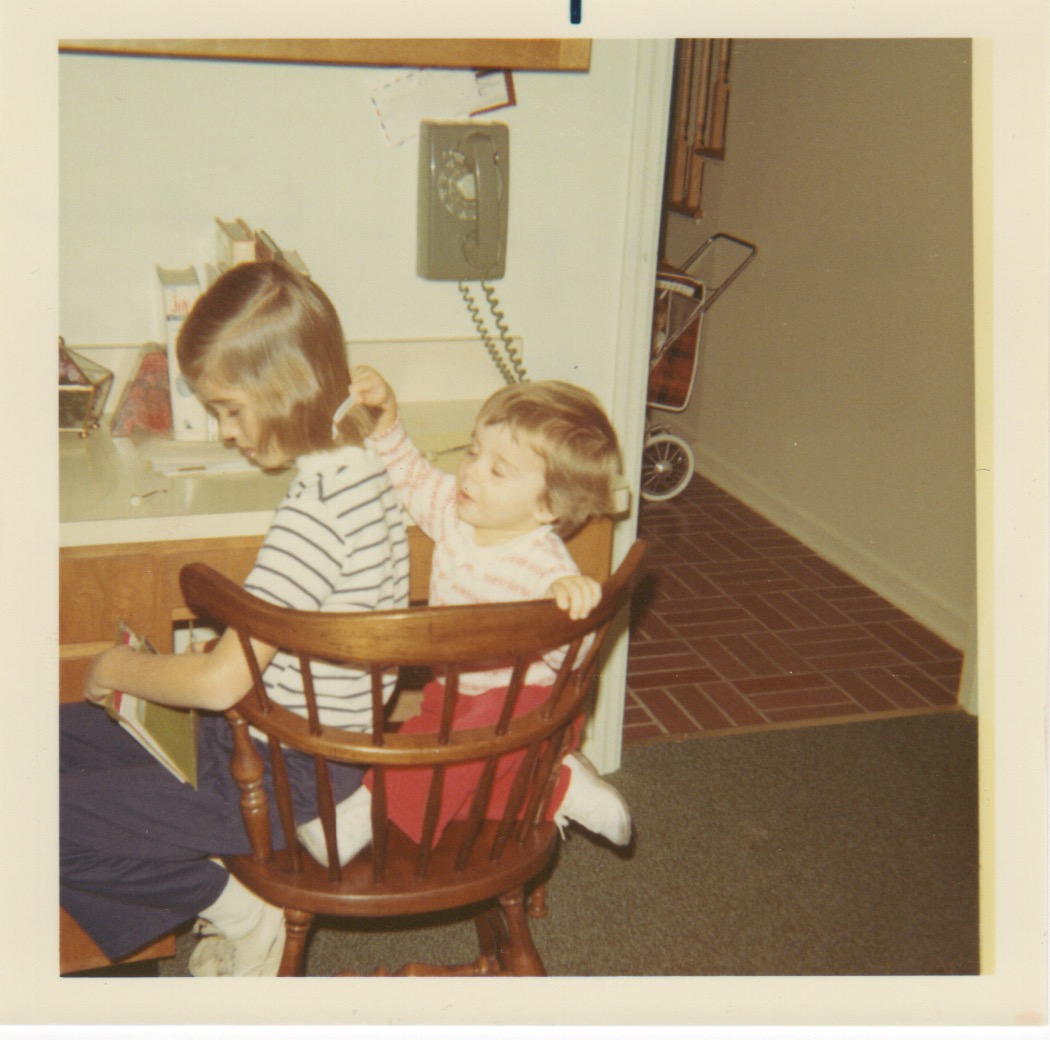 photo of Mary and big sister Pam. Photo circa early 70's. Pam is sitting on colonial style wooden chair while Mary sits behind her combing her hair. In the backgroud is a avocado green rotary wall phone above a laminate kitchen desk.