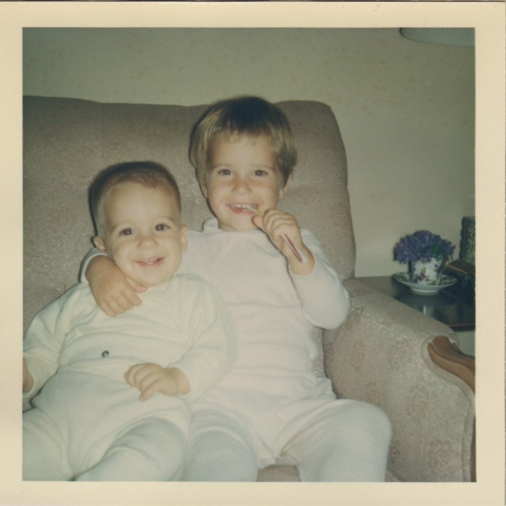 Pam and her "little" (now 6' 4") brother Pat sitting on a couch. This photo is circa early 60's. Pam has her arm around her brother and is chewing on a stras. Both children are dressed in white pajamas. There is a teacup on an end table to the side with purple flowers in it.